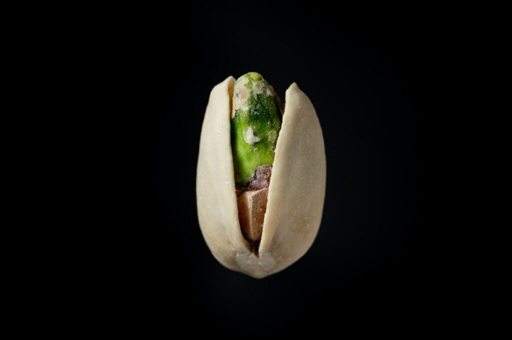 A single pistachio in an open shell on a black background. The vivid green nut contrasts against the pale beige shell, conveying simplicity.