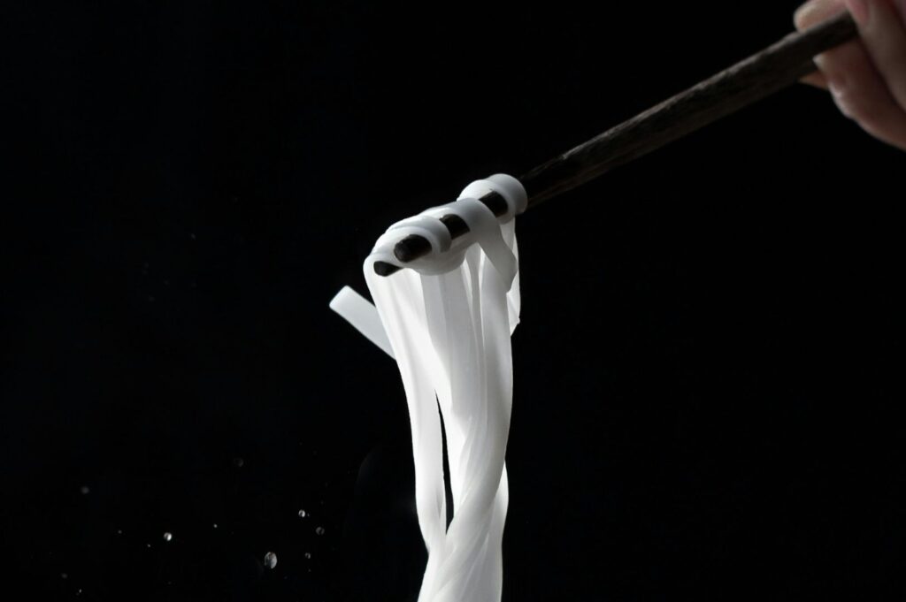 Chopsticks hold a swirl of white noodles against a dark background. The image is minimalist, highlighting the noodle texture and creating a serene, elegant tone.