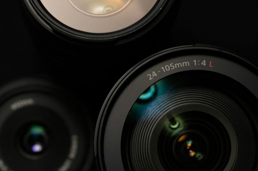 Close-up of three camera lenses against a dark background. The main lens in focus is labeled "24-105mm 1:4 L" and shows colorful reflections.