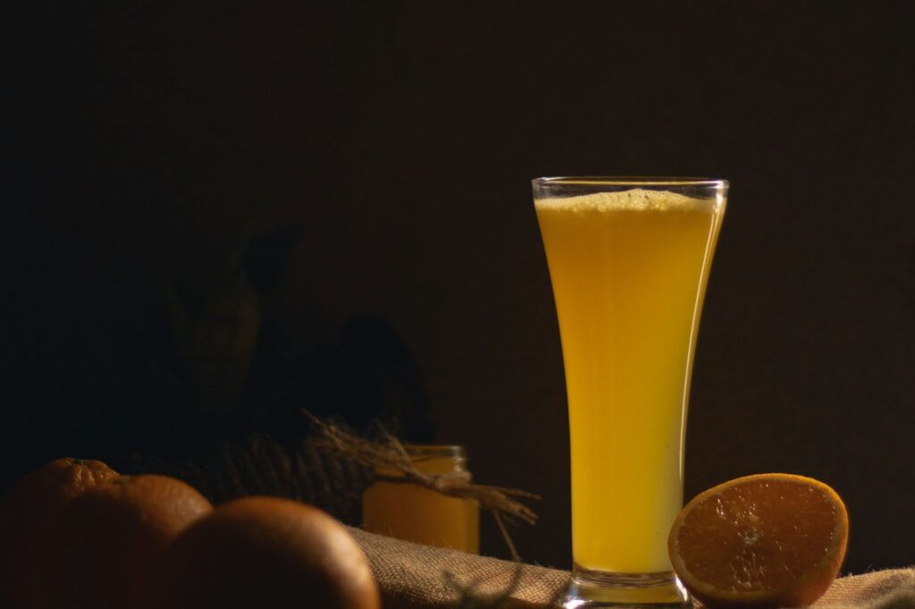 A tall glass of fresh orange juice sits on a table with a dark background, surrounded by whole and halved oranges, conveying a fresh and inviting tone.