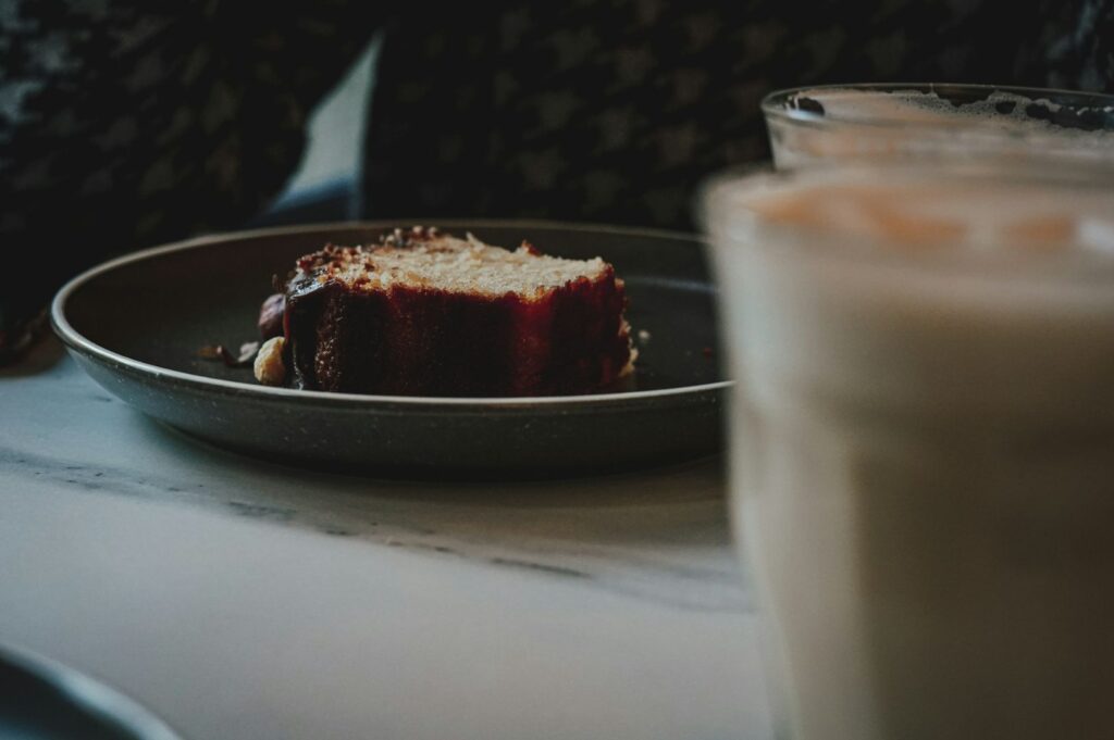 A slice of cake with a red glaze sits on a dark plate. In the blurred foreground, two frothy beverages are visible. The mood is cozy and inviting.