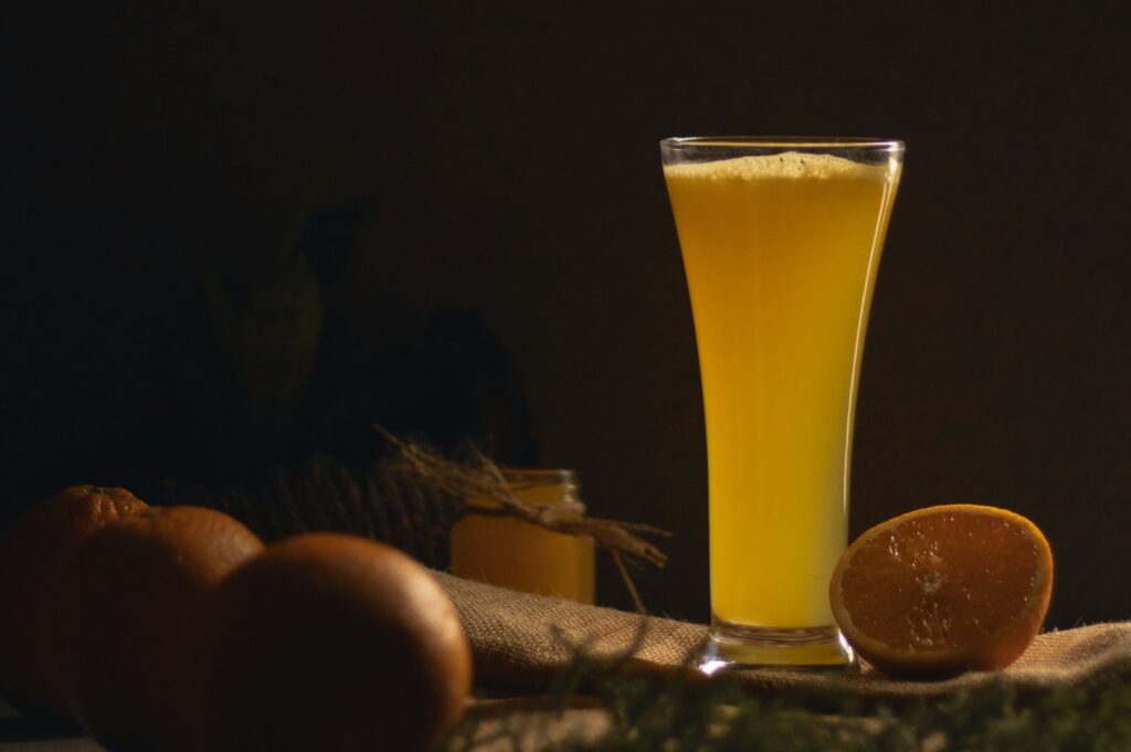A tall glass of frothy orange juice is set on a rustic table, accompanied by whole and halved oranges. The dim lighting creates a warm, cozy atmosphere.