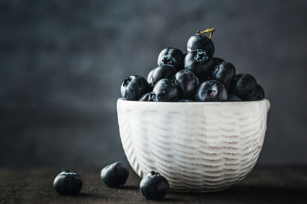 A white ceramic bowl overflowing with fresh blueberries sits on a dark surface. The moody background and soft lighting create a serene, inviting atmosphere.
