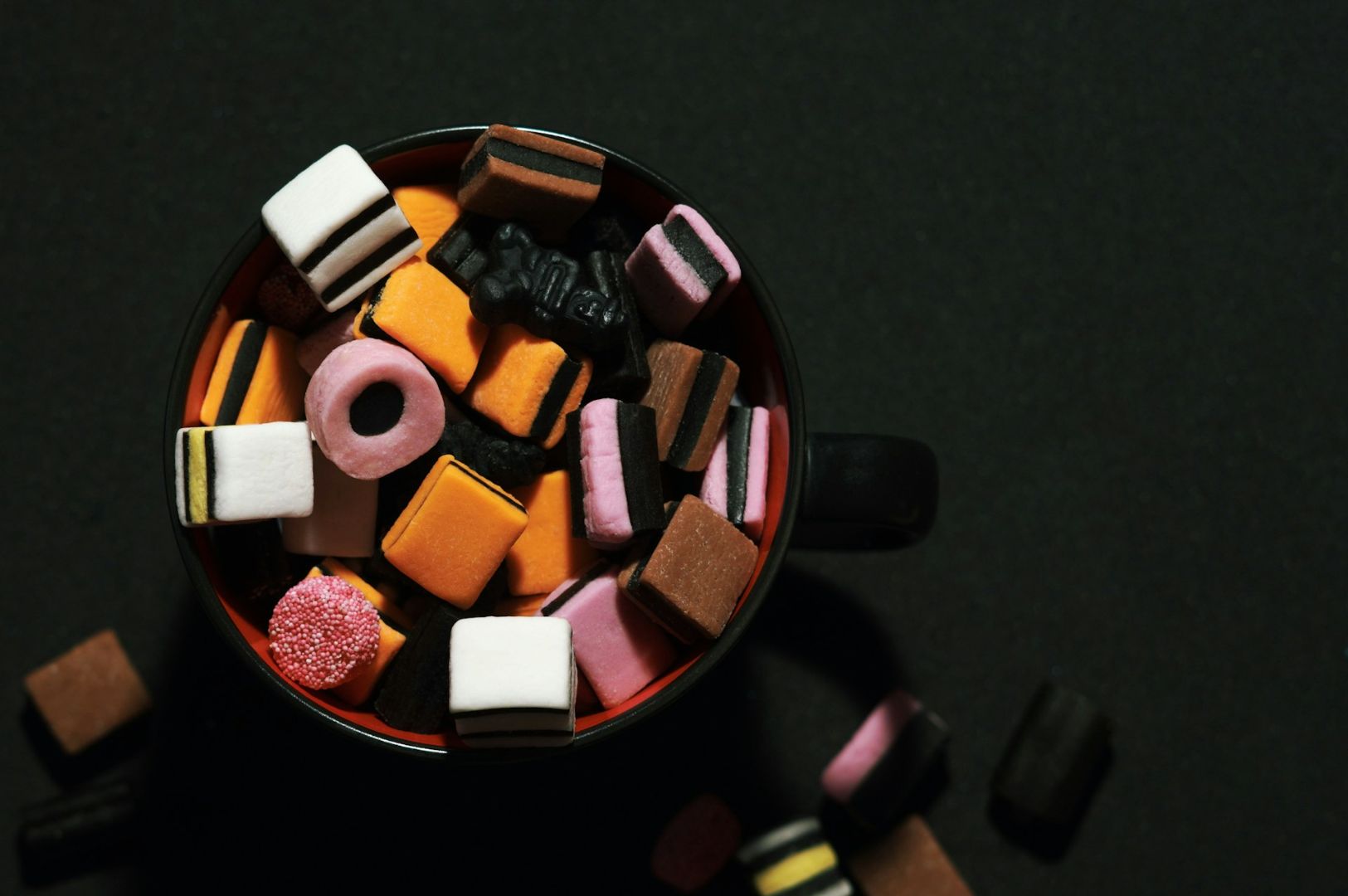 A cup filled with colorful licorice allsorts, including pink, orange, brown, and black pieces, sits on a dark background, conveying a playful tone.