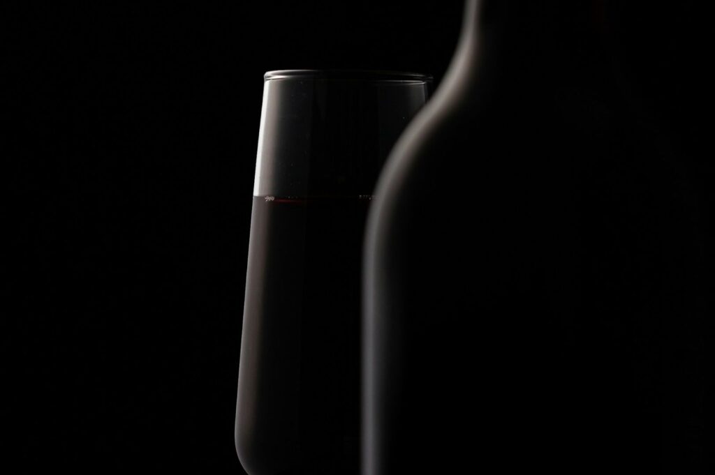 A wine glass half-filled with red wine stands beside an unfocused bottle, set against a dark background. The scene evokes a moody, elegant atmosphere.