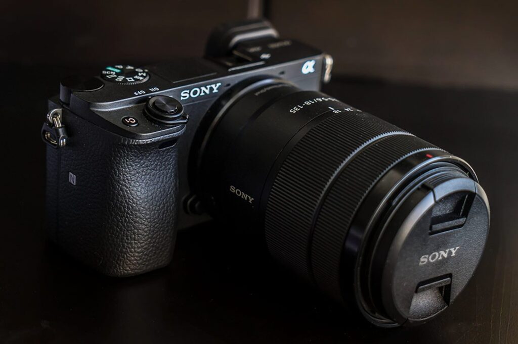 A black Sony mirrorless camera with a large zoom lens sits on a dark surface. The camera's textured grip and control dial are clearly visible.