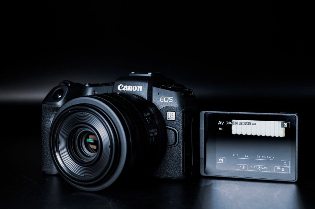 A Canon EOS camera with a lens on a dark background. The screen is flipped out showing settings in a minimalistic, professional setup.