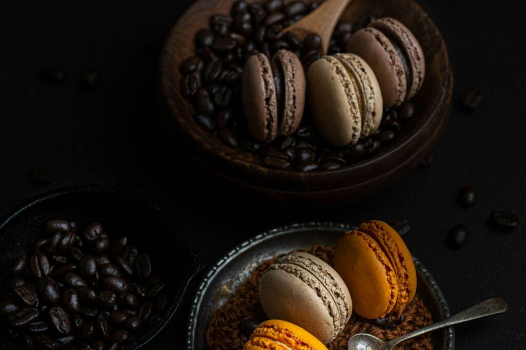 A bowl of coffee beans with chocolate macarons on top is paired with a metal plate holding orange and brown macarons, creating a cozy, elegant vibe.