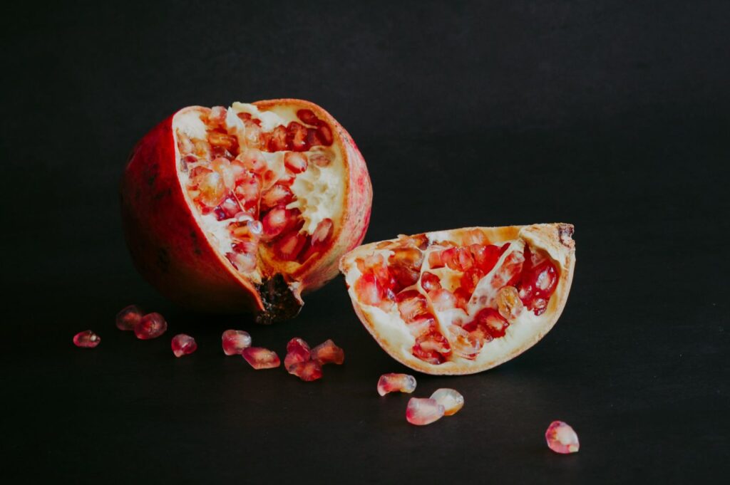 A halved pomegranate with glistening red seeds sits on a dark surface, surrounded by scattered seeds, creating a striking, fresh contrast.