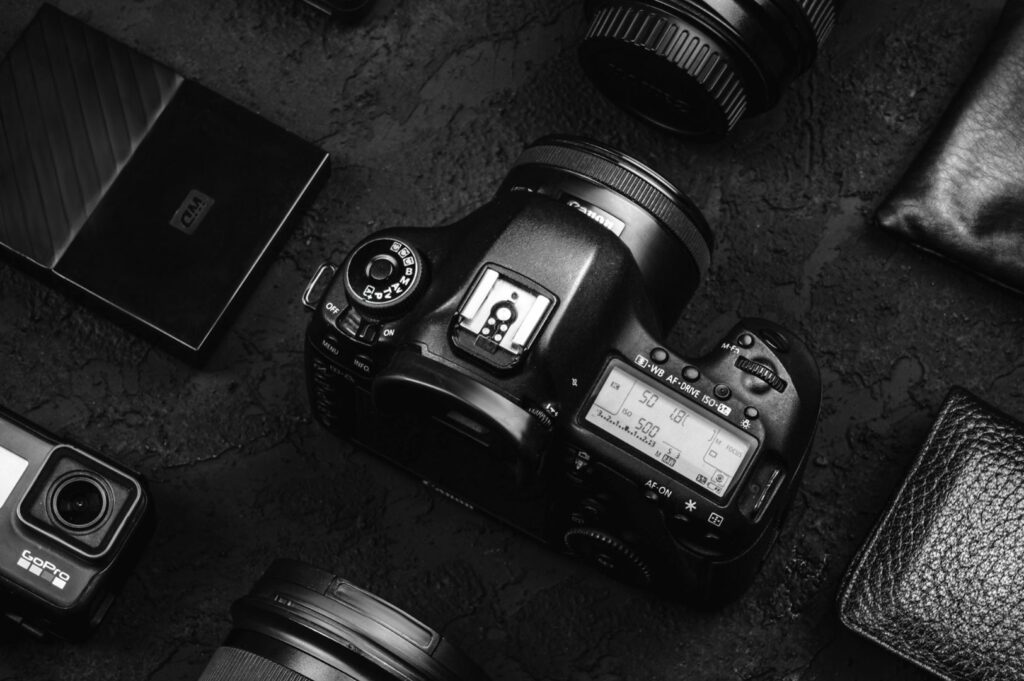 Black and white photo of a DSLR camera surrounded by lenses, a GoPro, a hard drive, and a wallet on a dark textured surface. Stylish and tech-focused.