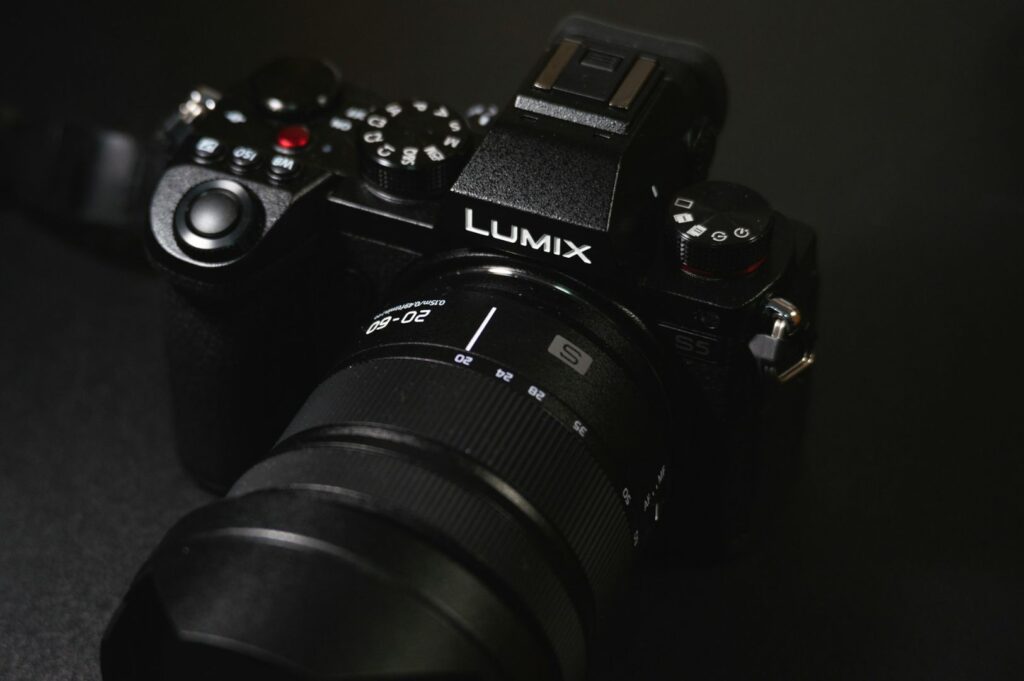 Close-up of a black Lumix camera with a zoom lens on a dark surface. The camera features multiple control dials and a prominent brand logo.
