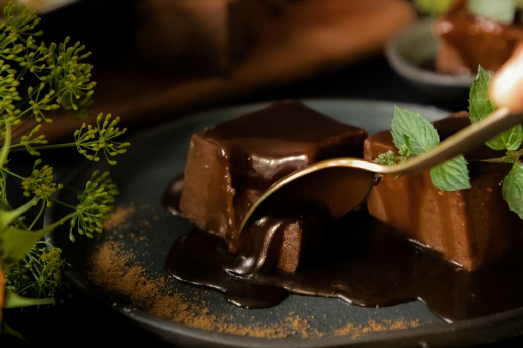 A fork cuts into a rich, glossy chocolate dessert square topped with mint leaves on a dark plate. Fresh green herbs and a sprinkle of cocoa surround it.