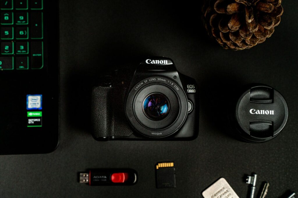 Flat lay of a Canon camera on a black surface, flanked by a lens cap, USB drive, SD card, laptop corner, and a pinecone, conveying a tech vibe.
