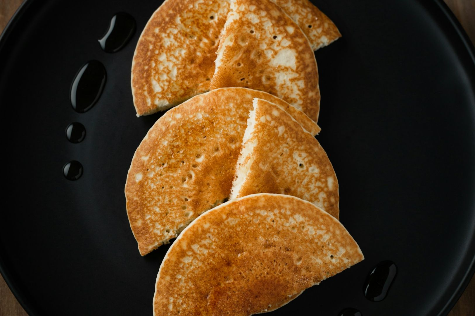 Golden brown pancakes cut into halves are artfully arranged on a black plate with syrup drizzles. The image evokes a feeling of warmth and indulgence.