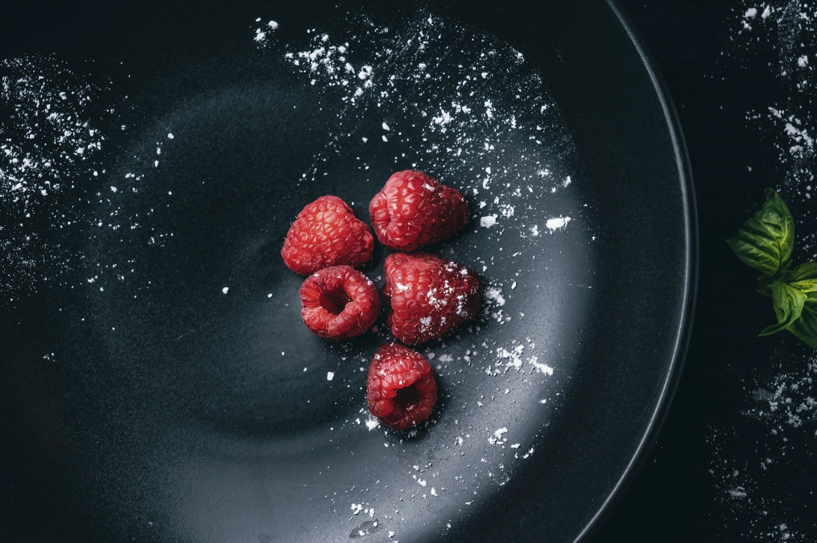 Four fresh raspberries with powdered sugar are arranged on a dark plate, creating a contrast. Fresh mint leaves are visible on the side, conveying a fresh, appetizing tone.