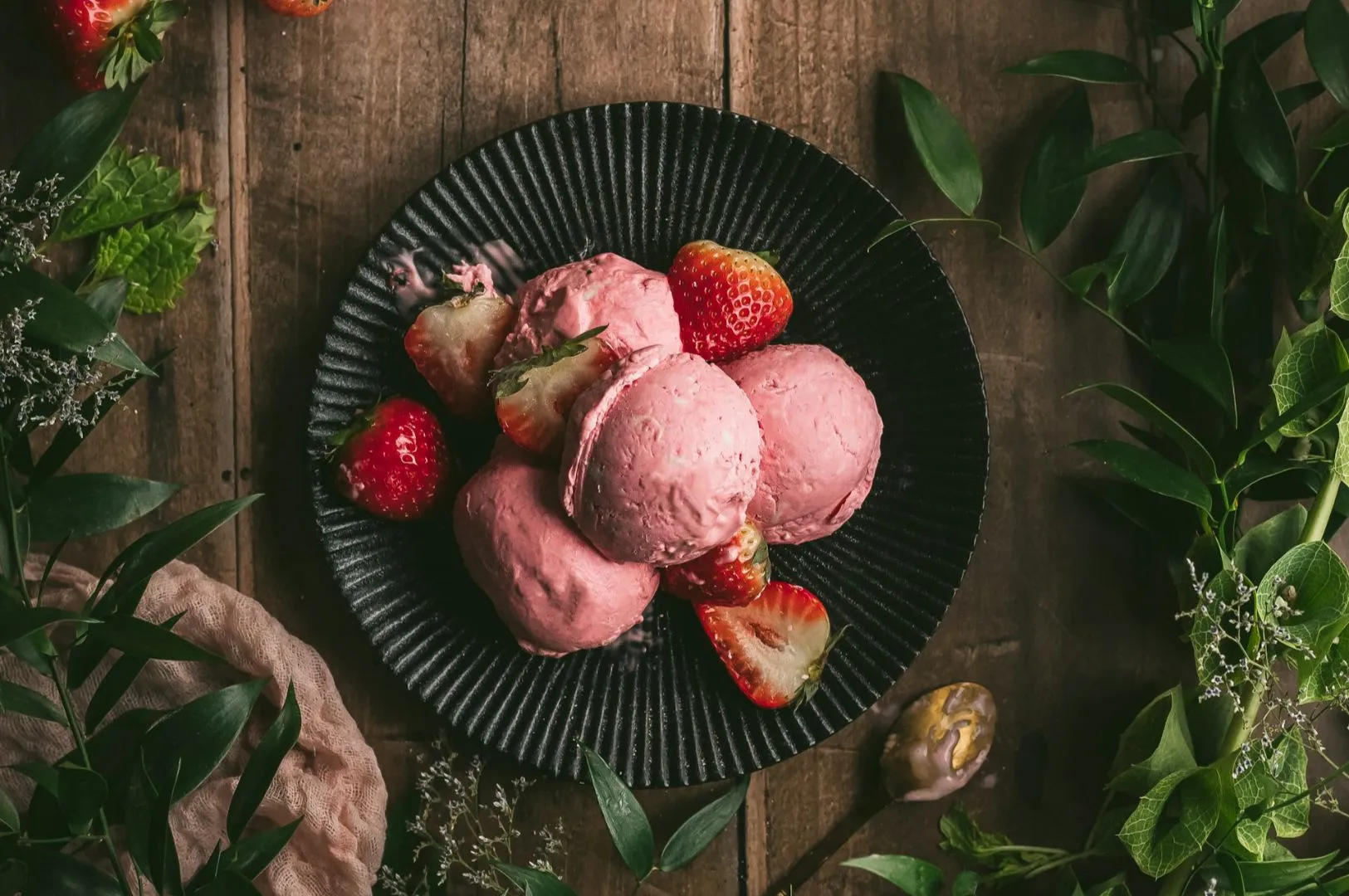 A black plate with scoops of pink strawberry ice cream and fresh strawberries, placed on a rustic wooden table, surrounded by green leaves and soft lighting.