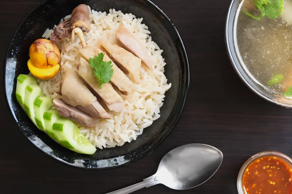 A black bowl holds sliced chicken on white rice, garnished with cilantro, accompanied by cucumber, and a sauce. A soup bowl and spoon are nearby on a dark table.