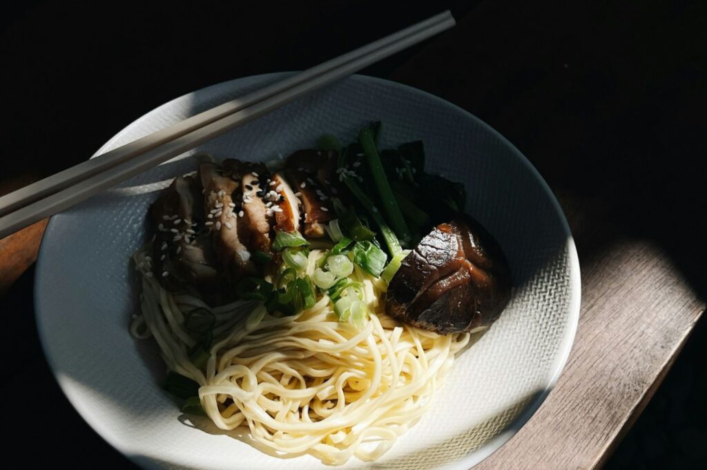 White ceramic bowl holds pale ramen noodles topped with slices of roasted pork, chopped green onions, and sesame seeds, with chopsticks laid across.