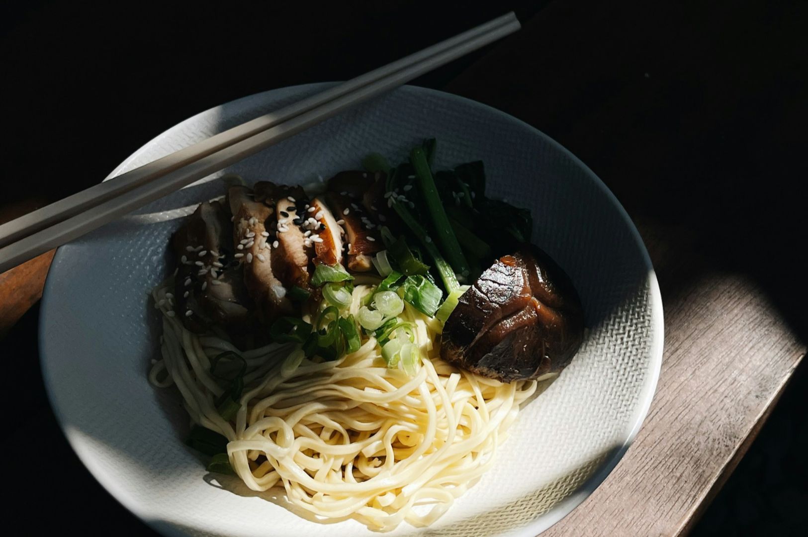 White ceramic bowl holds pale ramen noodles topped with slices of roasted pork, chopped green onions, and sesame seeds, with chopsticks laid across.