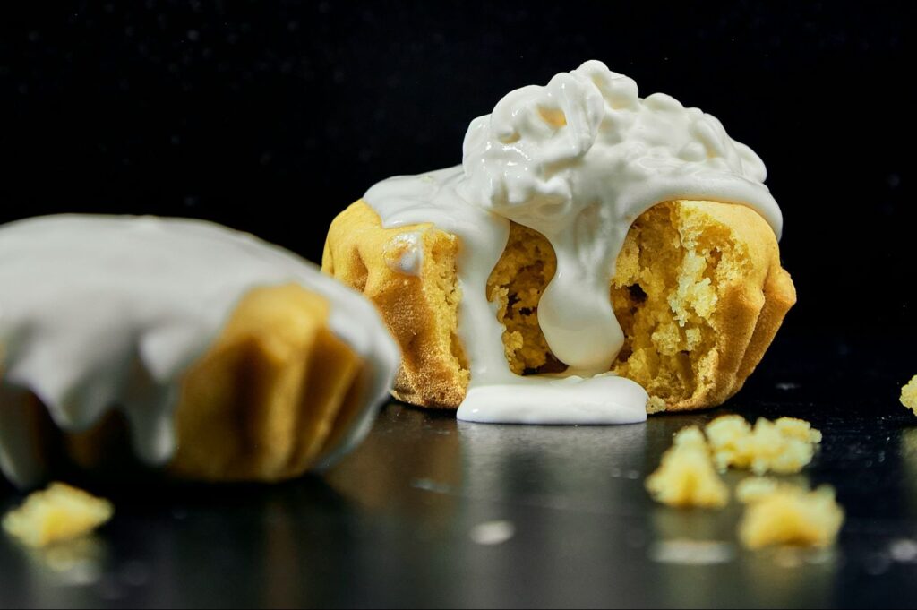 A muffin with a creamy white frosting overflows from the center, surrounded by crumbs on a dark surface, conveying a sense of indulgent delight.