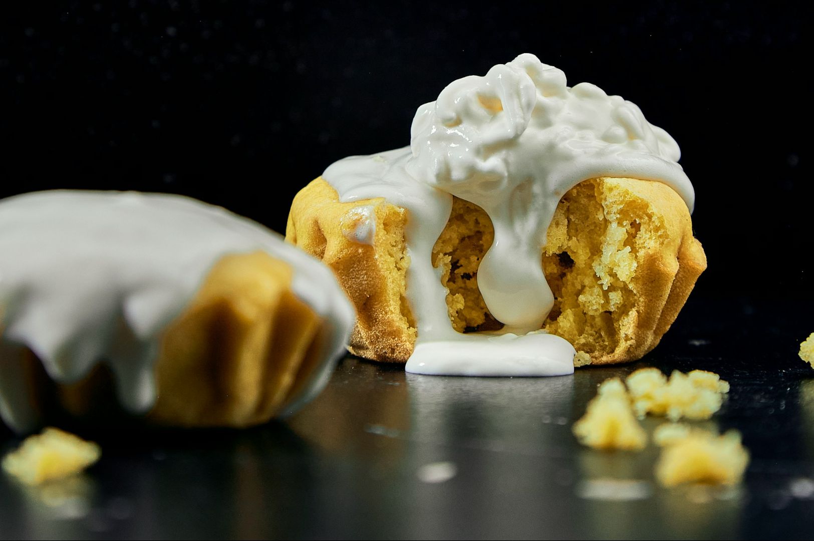 A muffin with a creamy white frosting overflows from the center, surrounded by crumbs on a dark surface, conveying a sense of indulgent delight.