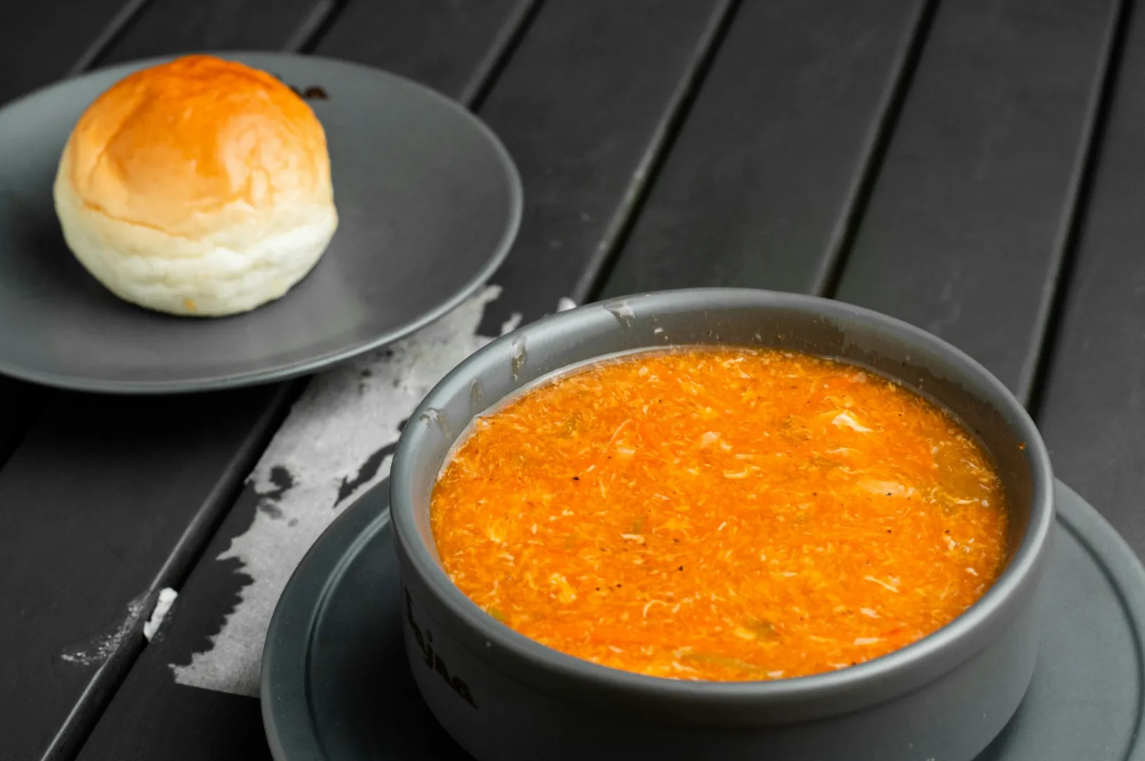 A bowl of vibrant orange soup sits on a dark table accompanied by a fluffy, golden bread roll. The scene evokes warmth and comfort.