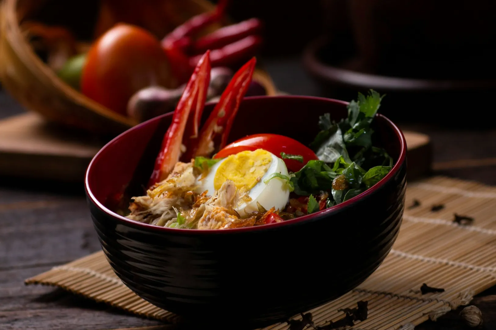 A bowl of ramen with shredded chicken, sliced red chilies, half a boiled egg, and fresh greens. The mood is warm and inviting, with rustic wooden tones.