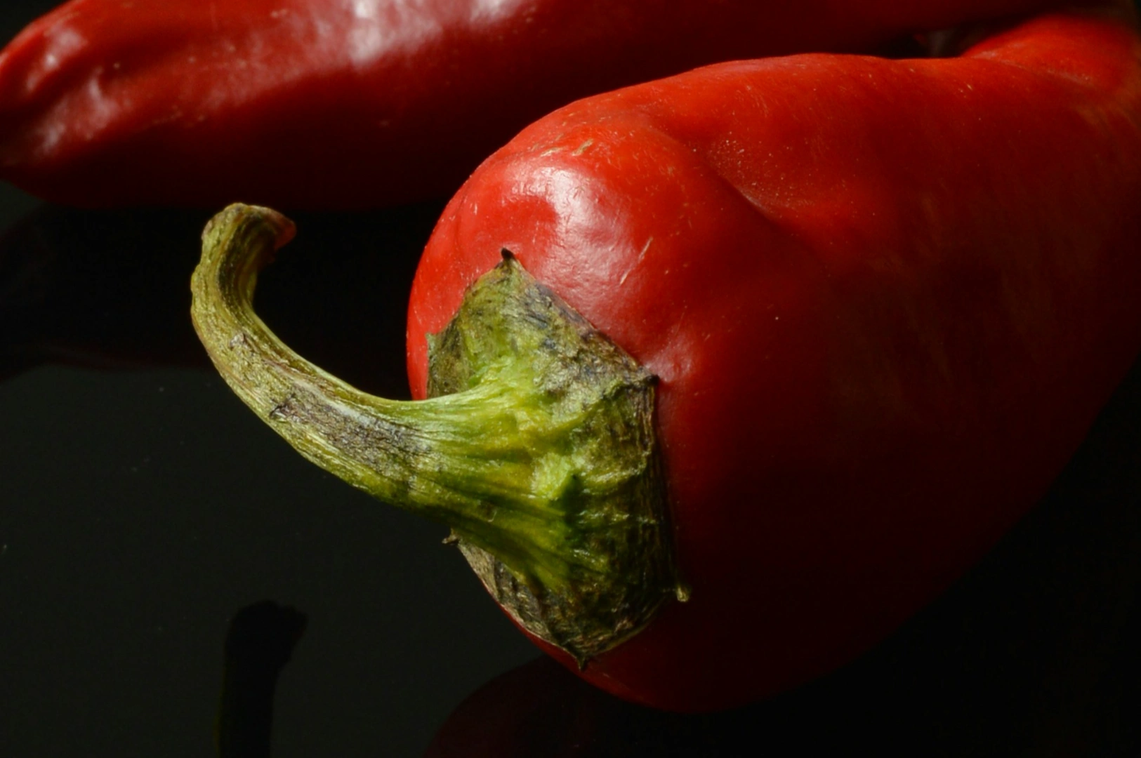 Close-up of a vibrant red bell pepper on a dark surface, showcasing its shiny skin and green stem. The lighting highlights its freshness.
