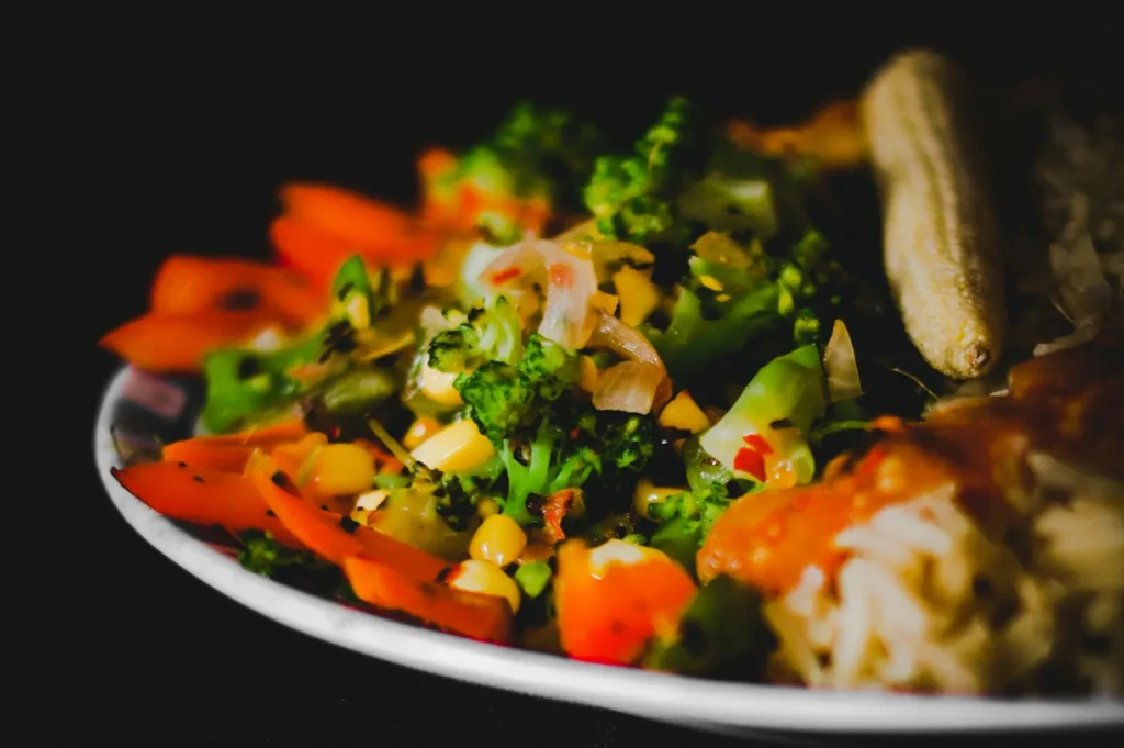 A vibrant plate of stir-fried vegetables, including broccoli, carrots, and corn, is artfully arranged with a side of banana, set against a dark background.