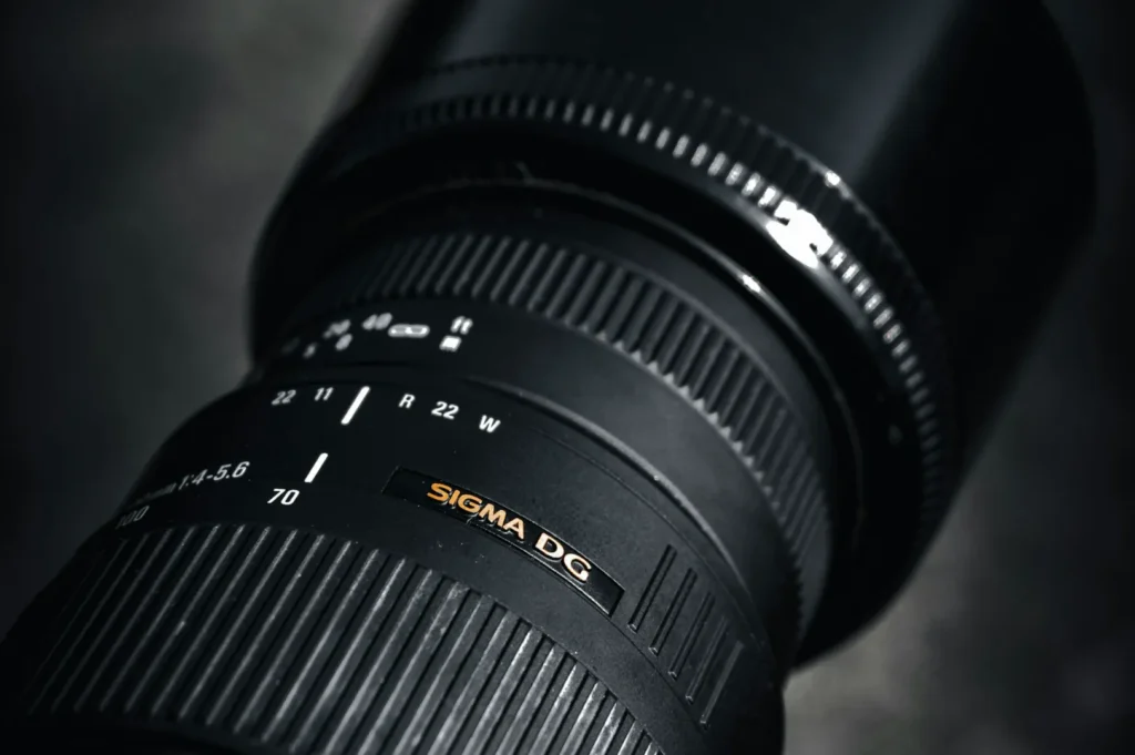 Close-up of a black Sigma DG camera lens with focus and zoom rings. The lens features white markings, conveying a sleek and professional tone.