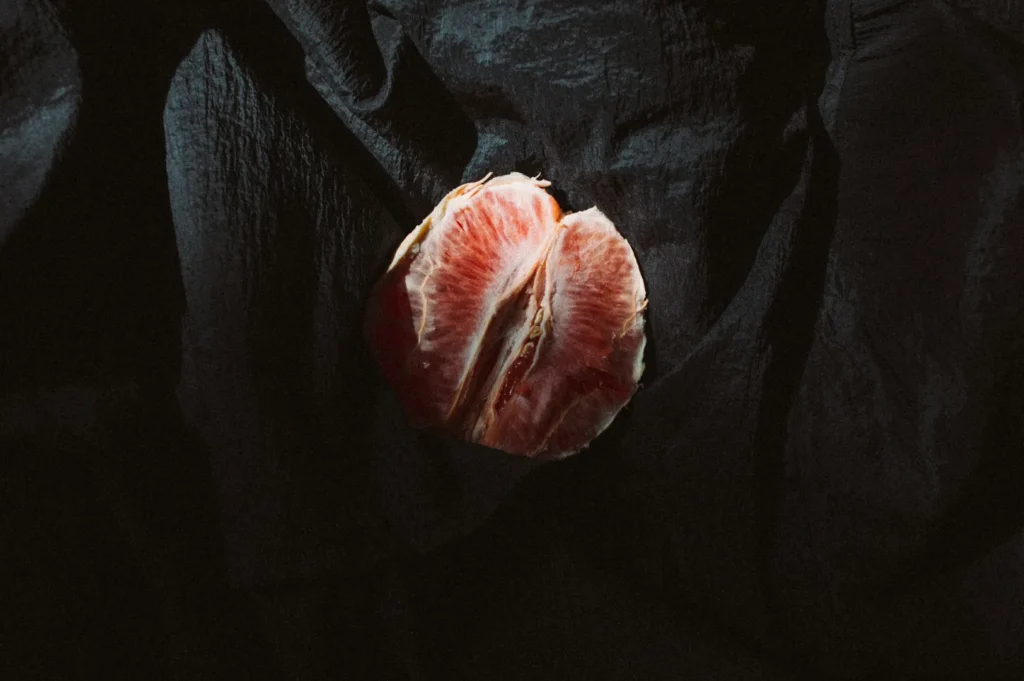 A halved grapefruit with vibrant pink flesh is placed on dark, textured fabric. The contrast creates a dramatic and moody atmosphere.