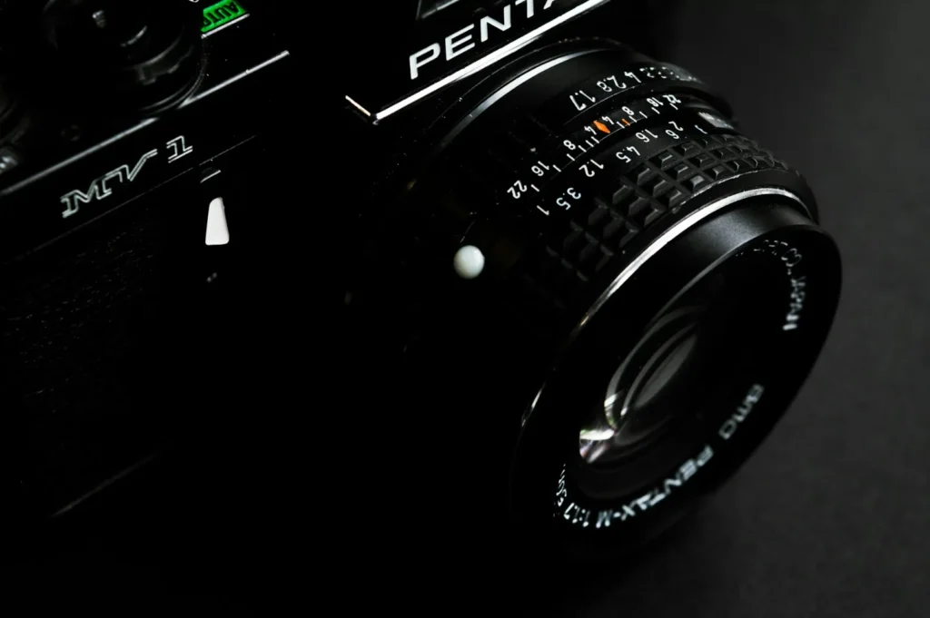 Close-up of a vintage Pentax MV1 camera lens on a dark background. The black and metal elements give a classic, nostalgic feeling.