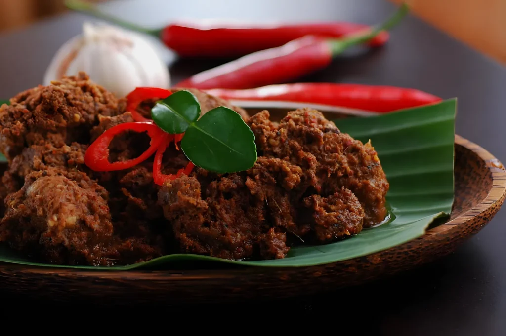 A serving of beef rendang on a banana leaf-lined plate, garnished with red chili slices and green leaves. Red chilies and garlic bulb in the background.