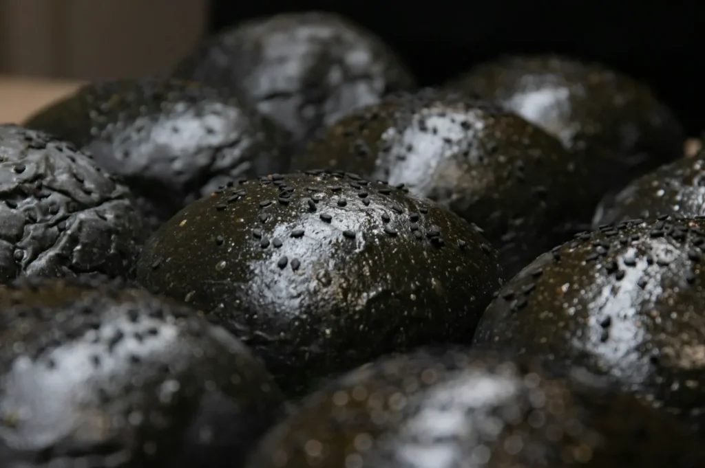 “Close-up of glossy black hamburger buns topped with black sesame seeds. The buns are tightly packed, creating a rich, dense texture.”