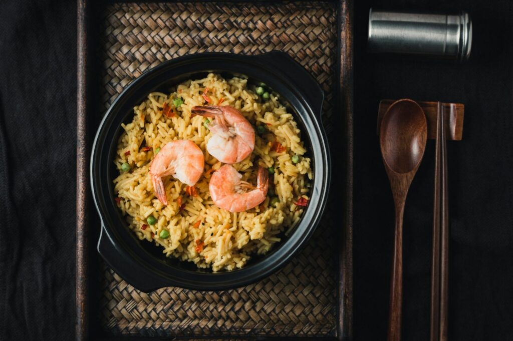 Top-down view of shrimp fried rice with plump seafood on the surface, colorful vegetables and fluffy grains, set against a dark background with a wooden spoon and chopsticks nearby.