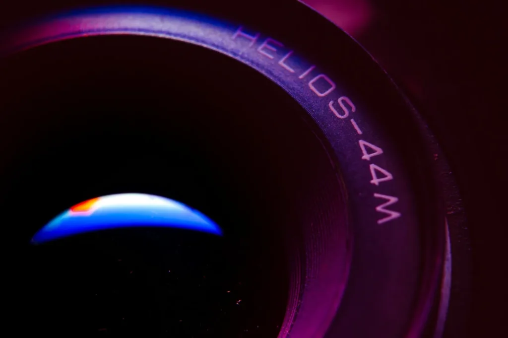 Close-up of a Helios-44M camera lens, with "Helios-44M" text visible. The lens reflects a subtle blue and orange light against a dark, moody background.
