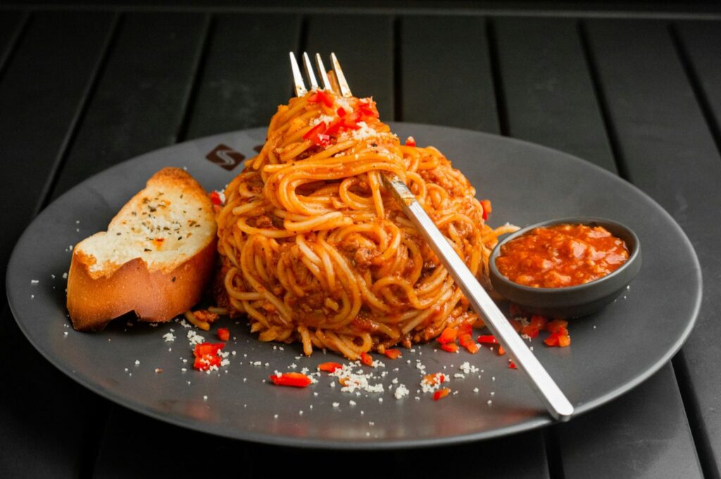 A plate of spaghetti bolognese garnished with red peppers and grated cheese is centered on a dark plate. A fork is woven into the pasta. To the side, there is a slice of garlic bread and a small dish of red sauce. The setting is elegant with a dark-toned background.