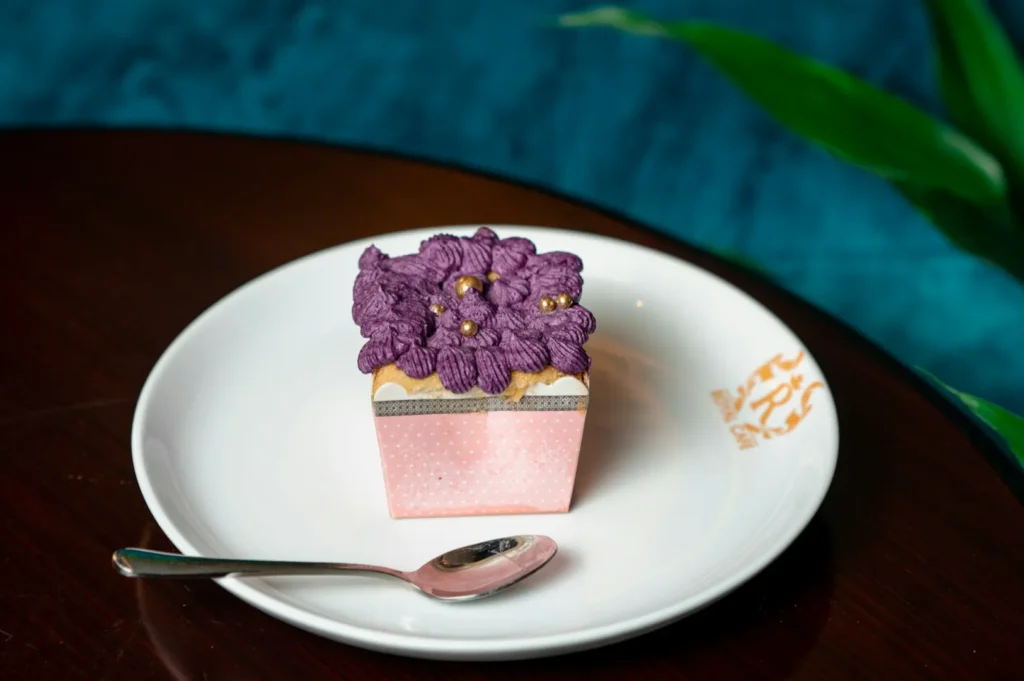 A pink cupcake on a white plate with intricate purple frosting resembling flowers and gold accents. A spoon lies beside it. Warm, elegant setting.