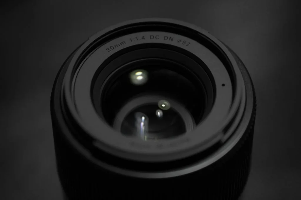 Close-up view of a camera lens with "30mm 1:1.4 DC DN Ø52" text. The reflective glass and dark background give a sleek, professional feel.