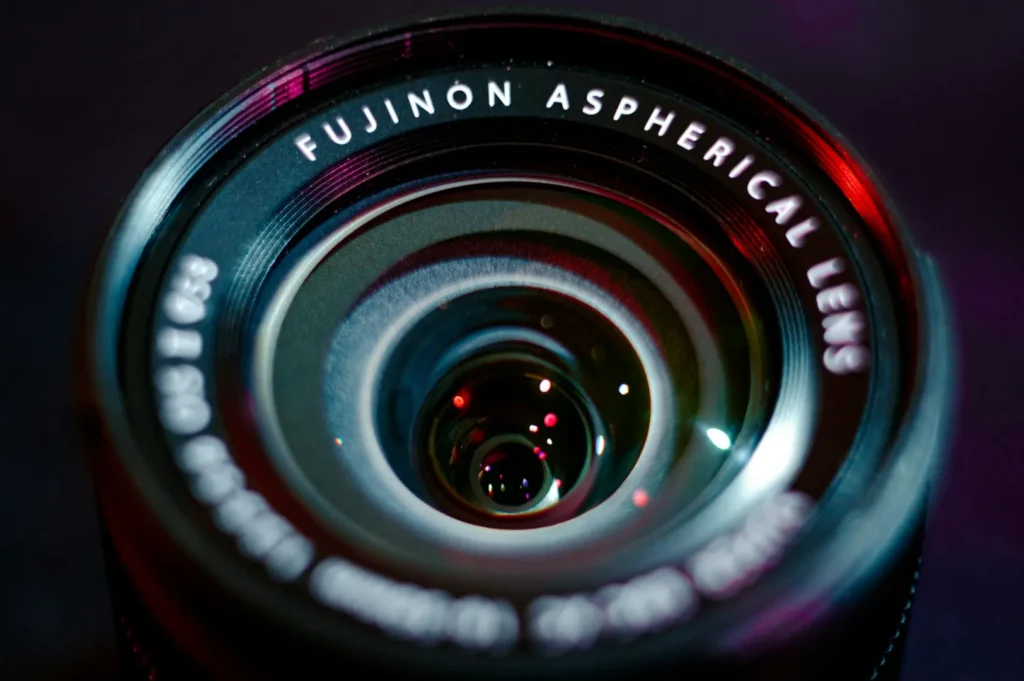 Close-up of a Fujinon aspherical camera lens, reflecting light in shades of red and blue. The lens details are sharp, creating a technical and focused mood.