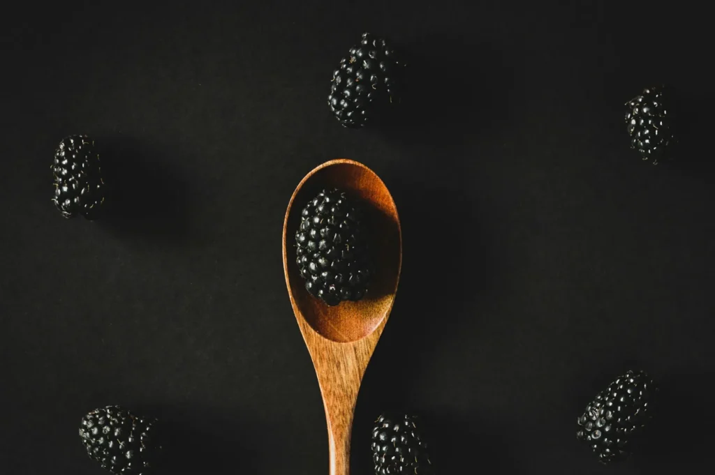 Wooden spoon holds a blackberry against a dark backdrop. Additional blackberries are scattered around, creating a minimalist, elegant contrast