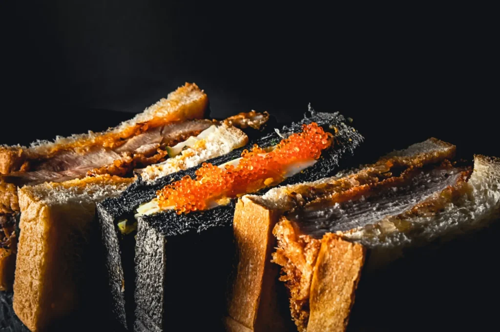 A variety of gourmet sandwiches are displayed against a dark background. Some have black bread with bright red roe, while others feature crispy, textured fillings.