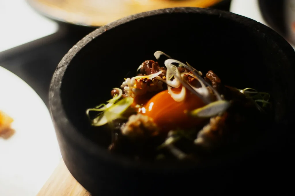 “A close-up of a dish in a black bowl featuring a raw egg yolk, garnished with thinly sliced spring onions and herbs, creating a rustic, gourmet vibe.”