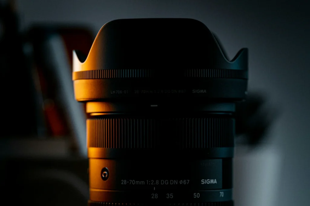 Close-up of a camera lens, partially illuminated by warm light, against a dark background. The lens detail and shadows create a moody atmosphere.