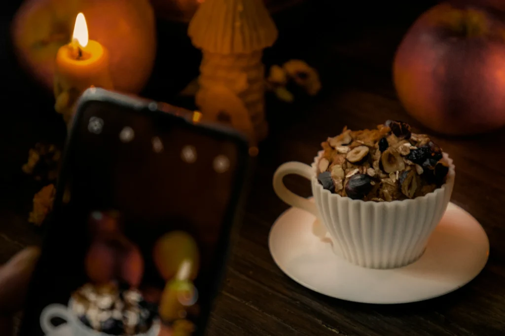 A dimly lit scene with a lit candle, an apple, and a white teacup holding granola. A smartphone is capturing the setup, creating a cozy, warm ambiance.