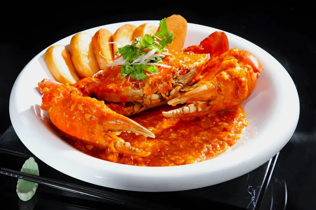 A white plate holds chili crab in vibrant red sauce, garnished with greens and served alongside toasted buns, creating an appetizing, spicy scene.