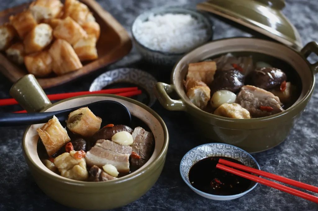 Two clay pots filled with braised pork, mushrooms, garlic, and tofu sit on a table. Accompanied by bowls of rice, dipping sauce, and a plate of crispy dough sticks. Red chopsticks add a touch of color, creating a cozy, inviting meal setting.