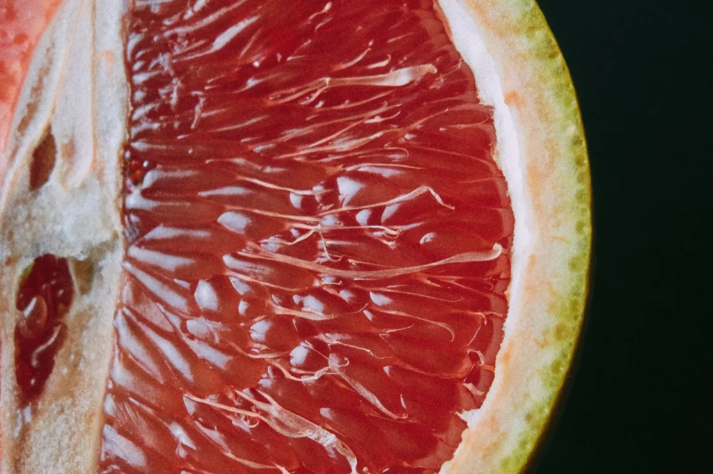 Close-up of a sliced grapefruit showing vibrant, juicy red pulp with glistening texture, surrounded by a yellowish rind on a dark background.
