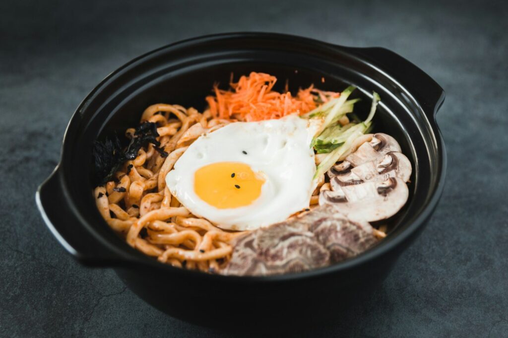 A black ceramic bowl holds bibimbap-styled noodles topped with a sunny-side fried egg, sautéed mushrooms, shredded carrot, cucumber strips, and seaweed, all arranged in a steaming, glossy presentation.