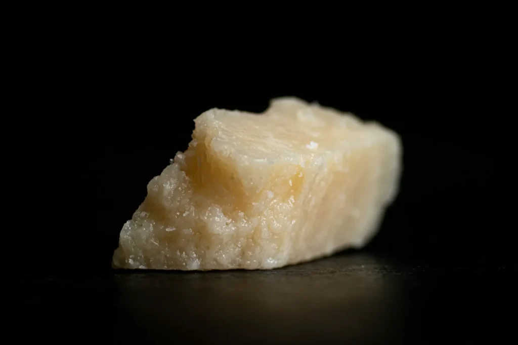 Close-up of a small, textured Parmesan cheese chunk on a dark background. The cheese is pale yellow, with a rough and grainy surface.